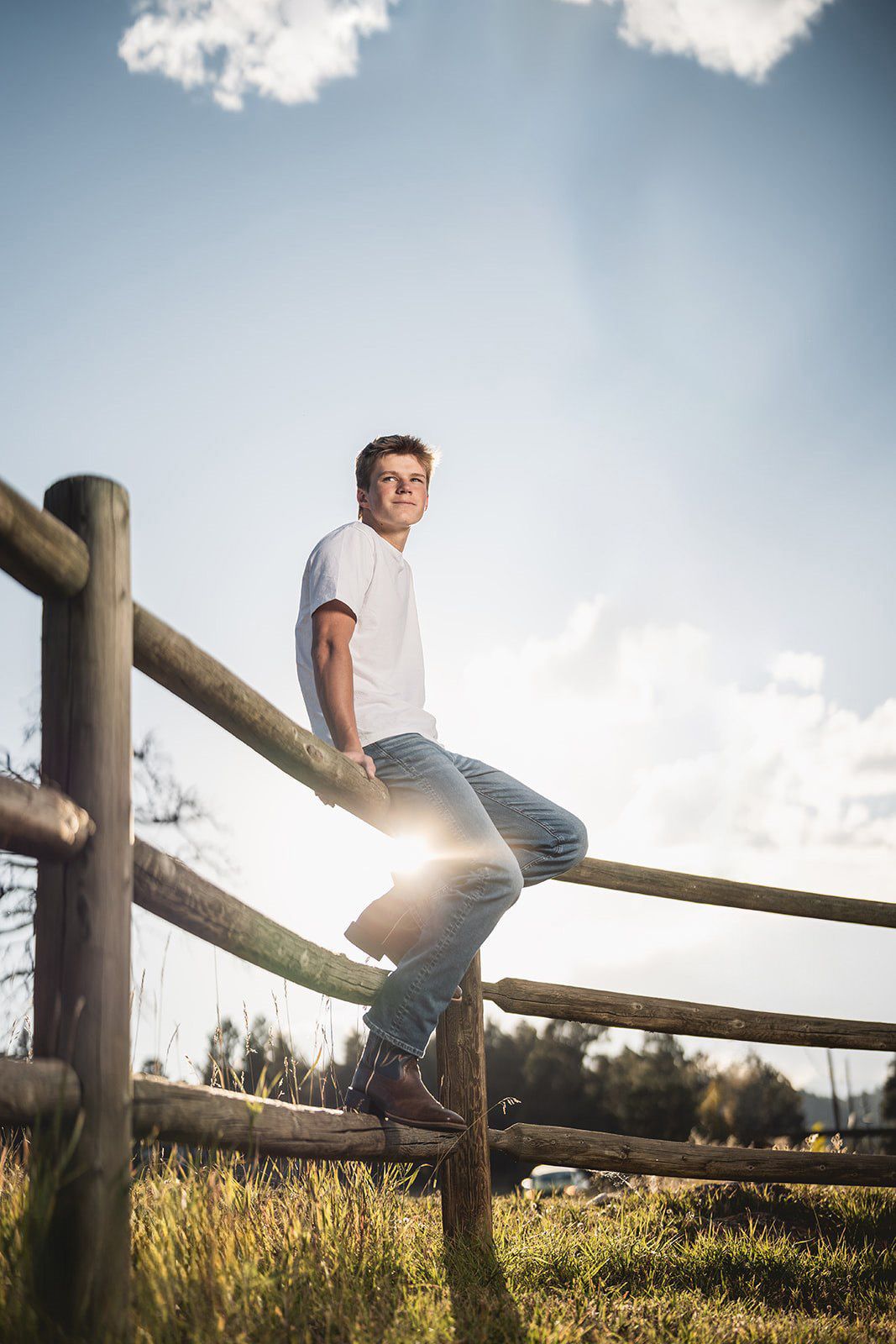 Person sitting on wooden fence rail against blue sky with fluffy clouds.