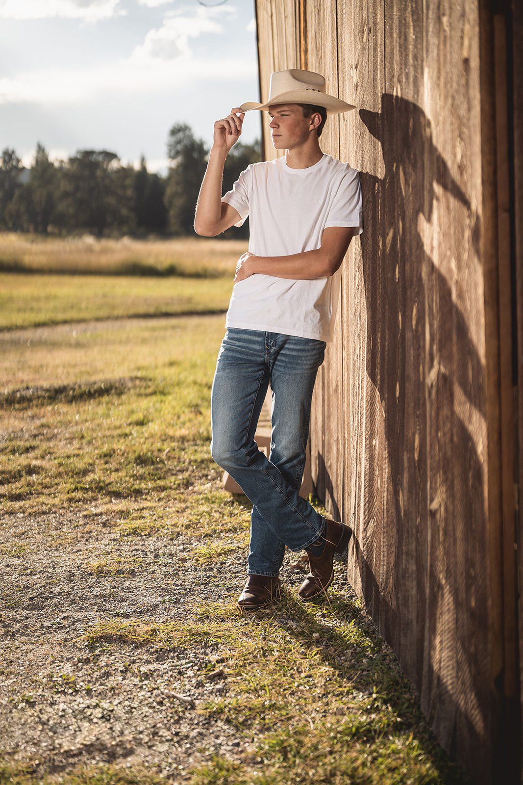 Cowboy in white tee and jeans tips hat while leaning on wooden barn exterior.