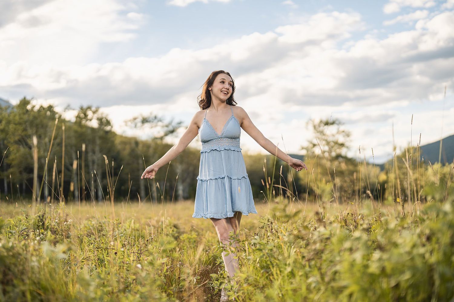 Smiling young woman in light blue sundress twirling in a wildflower meadow under cloudy skies.