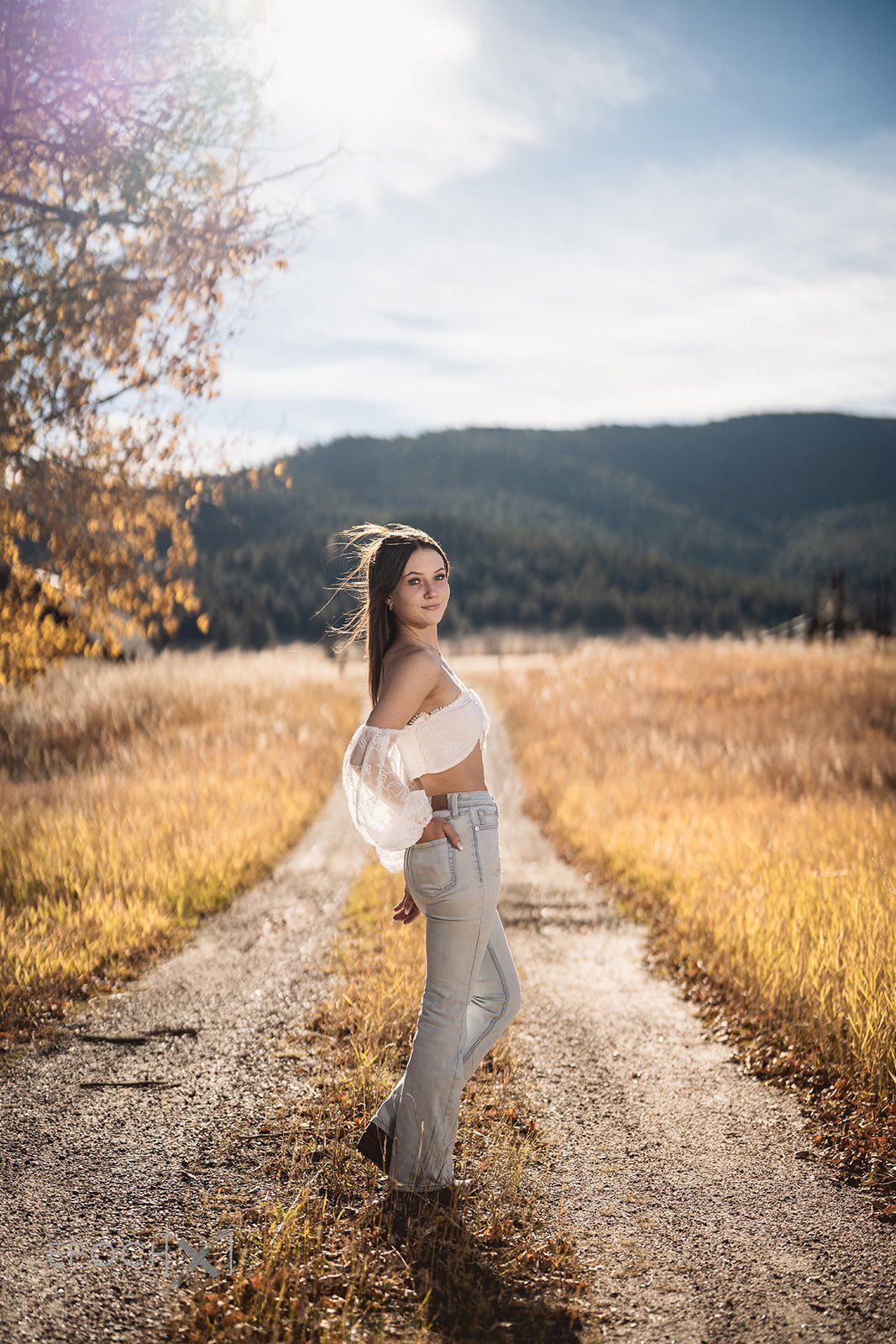 Young woman in white off-shoulder top and jeans standing on a rural dirt road with mountain backdrop.