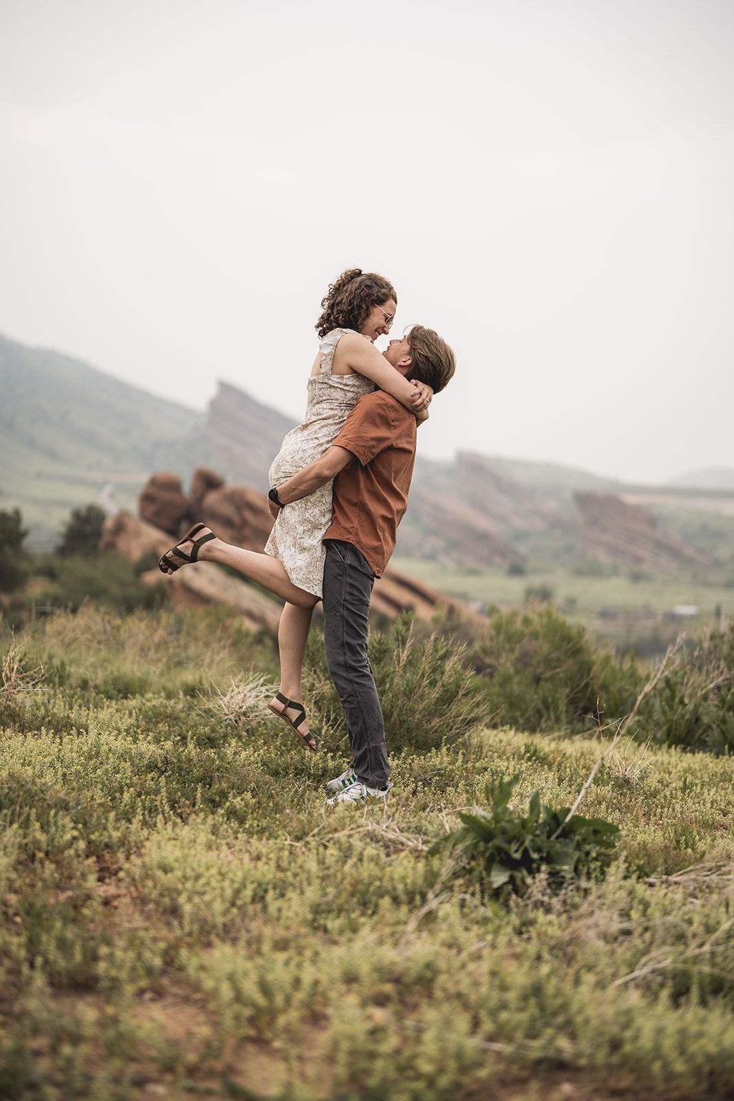 Man lifting woman in an embrace among green shrubs with red rock formations visible in the misty background.