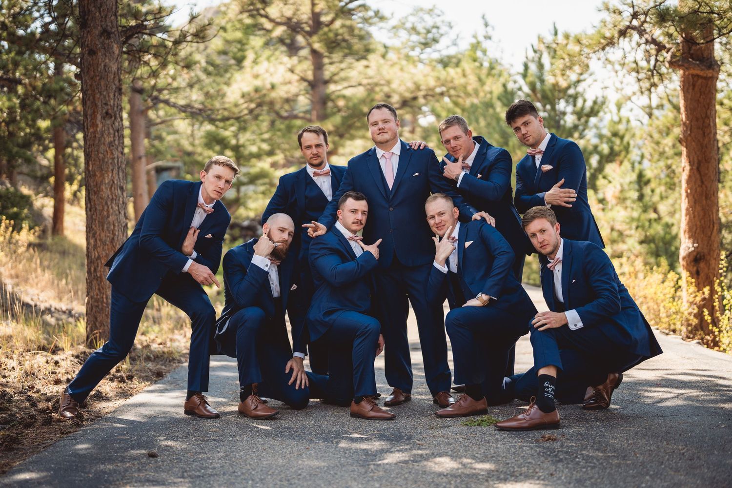 Groomsmen in navy blue suits pose playfully on a tree-lined path for a wedding party photo.