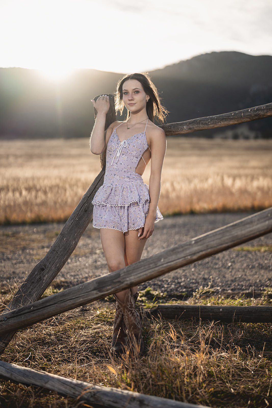 Young woman in a floral romper leaning on a rustic wooden fence at golden hour sunset.