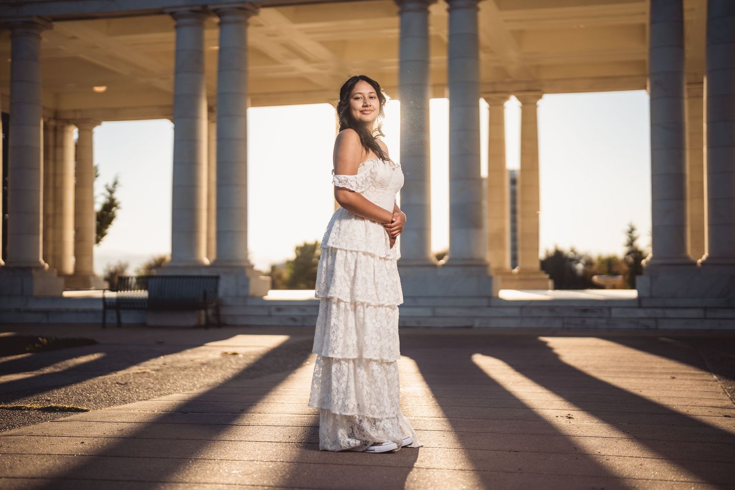 Woman in white tiered dress standing among stone columns in golden hour sunlight.
