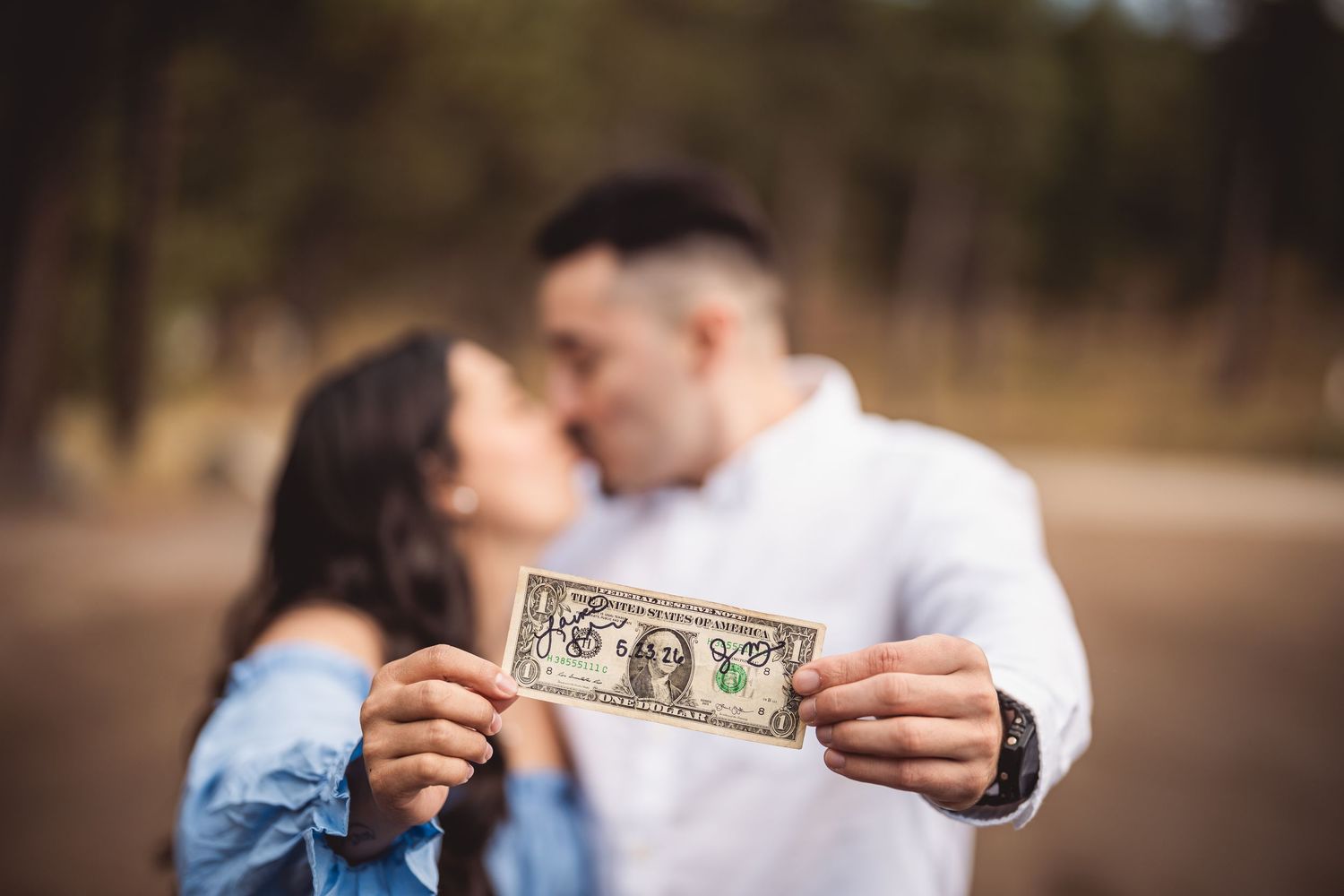 Couple kissing while holding a decorated one dollar bill in focus outdoors.