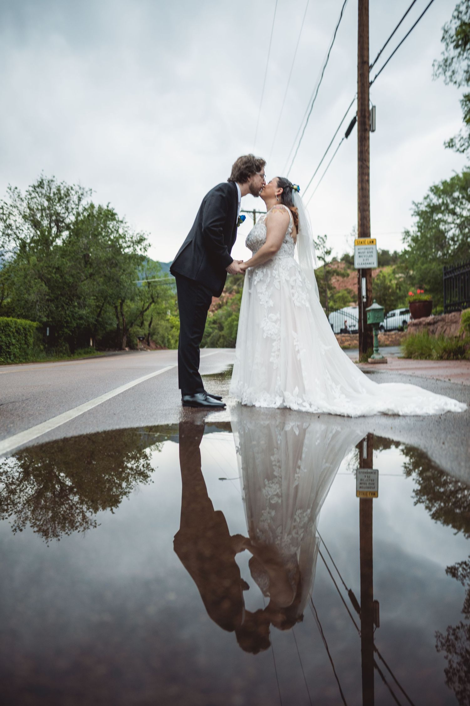 Wedding couple's reflection captured in rain puddle during overcast outdoor photoshoot.