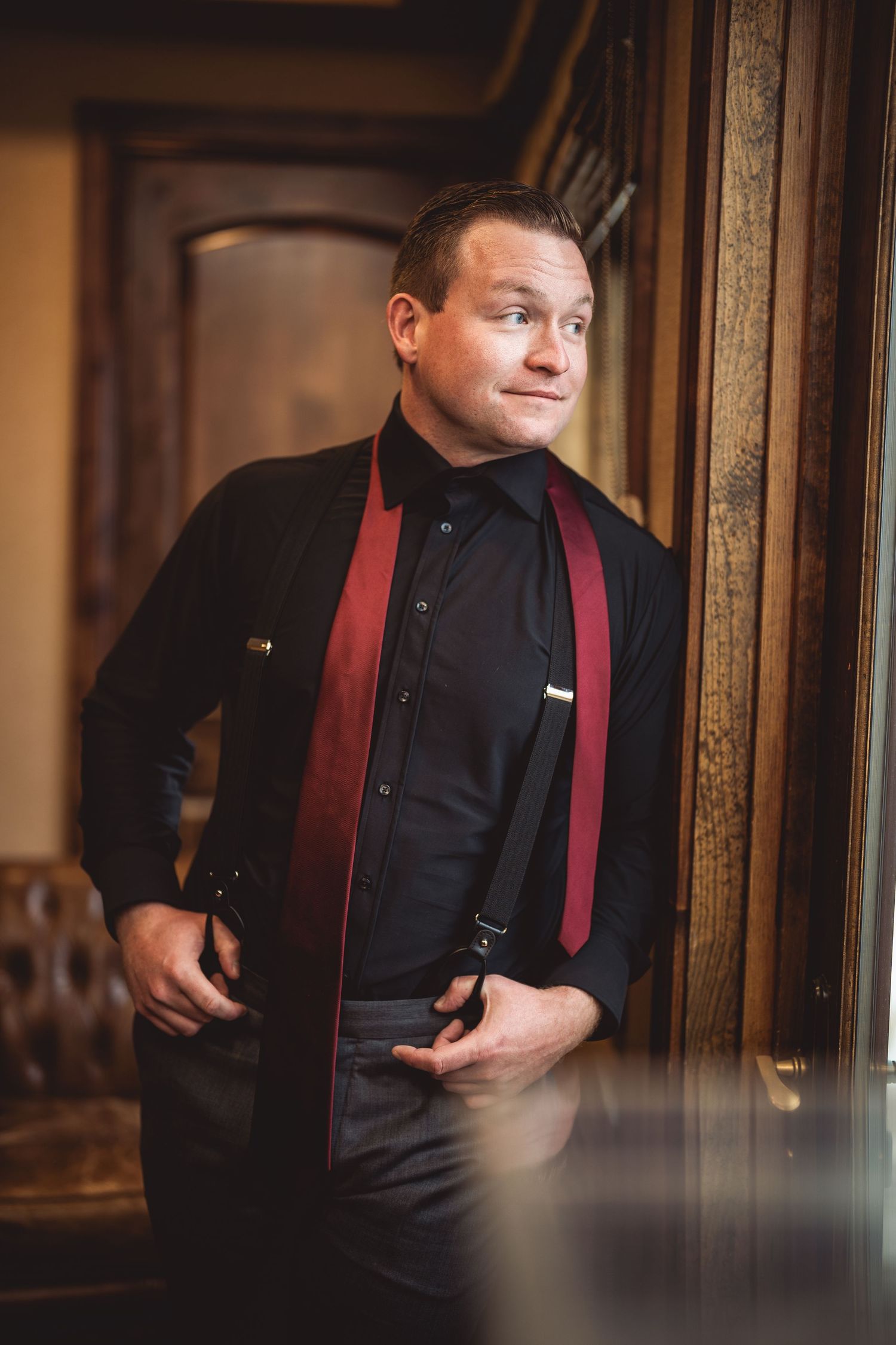 Person wearing a black suit with burgundy suspenders posing against an indoor backdrop.