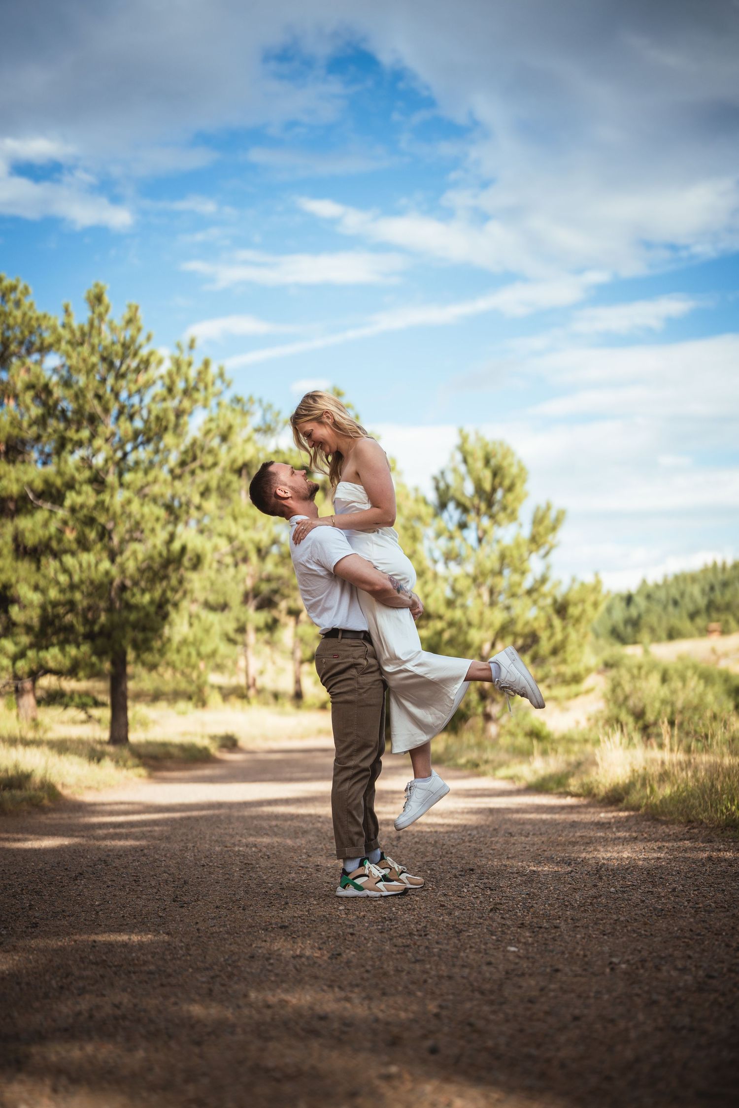 Man lifting woman in white dress on a scenic forest path, sharing a romantic moment under a blue sky.
