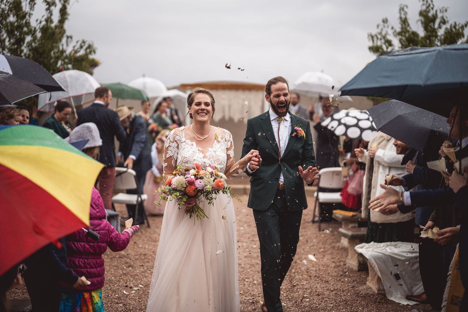 Rainy outdoor wedding ceremony with colorful umbrellas and standing guests.