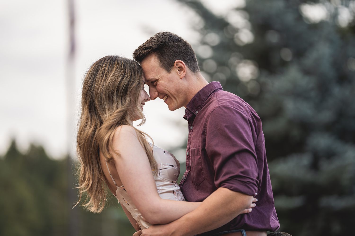 Couple sharing tender moment in front of evergreen trees during sunset.