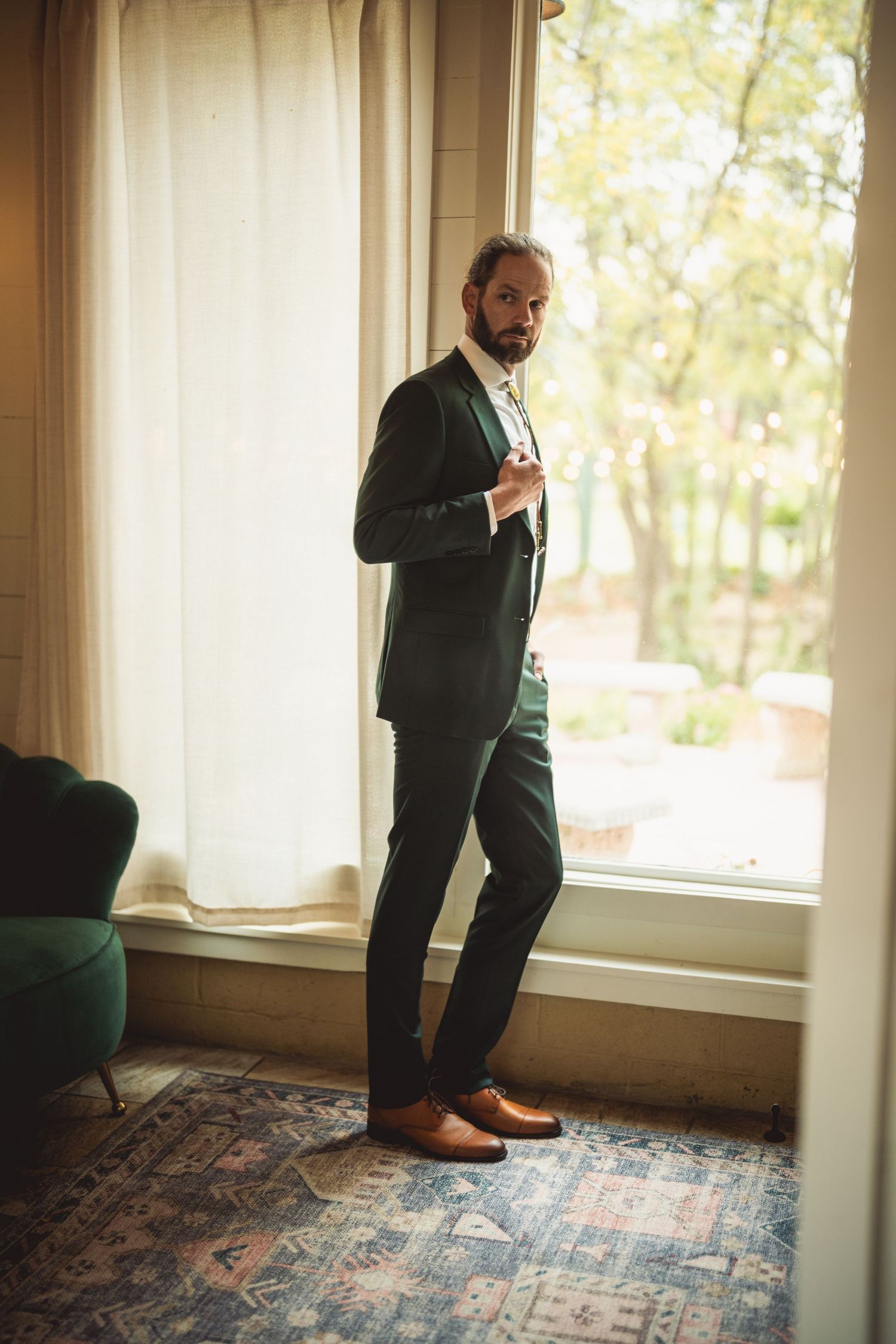 Groom in formal attire standing by bright windows on patterned carpet.