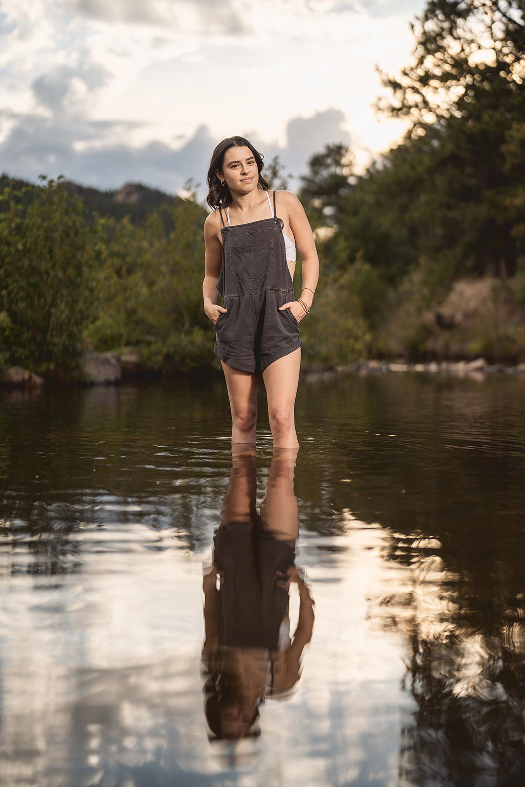 Woman stands in calm lake water during sunset with mountains in background.
