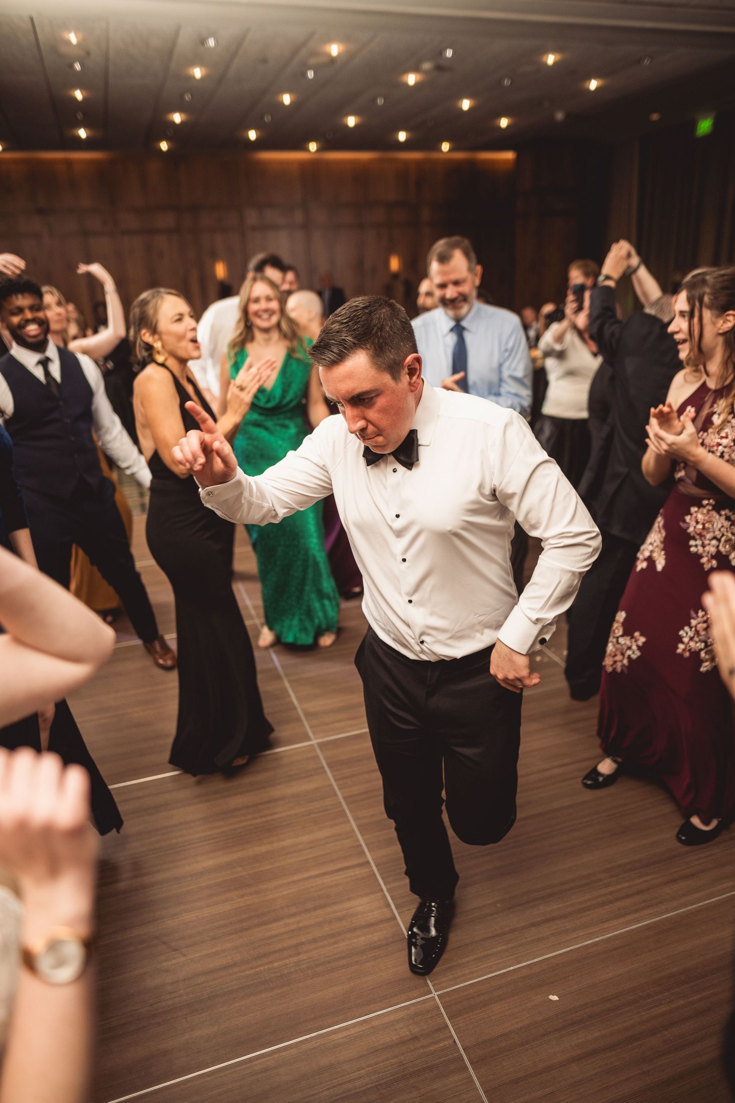 Wedding guests dance enthusiastically on a wooden dance floor under warm lighting.