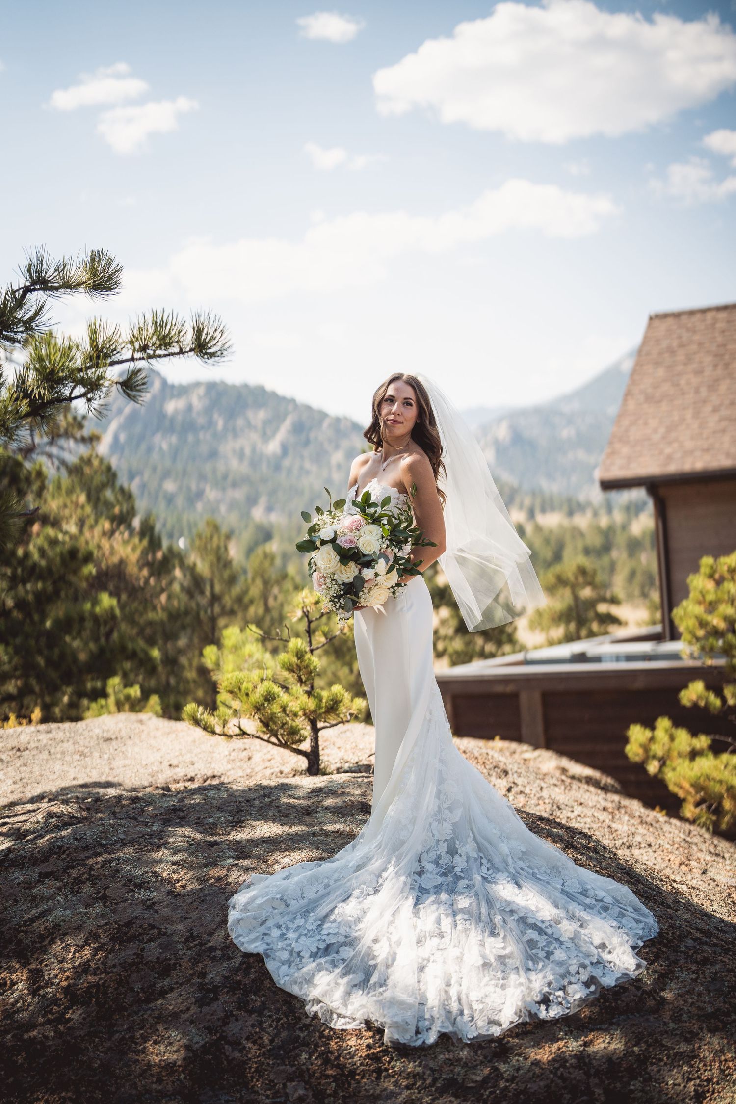 Bride in flowing lace gown poses against mountain backdrop with rustic cabin visible.