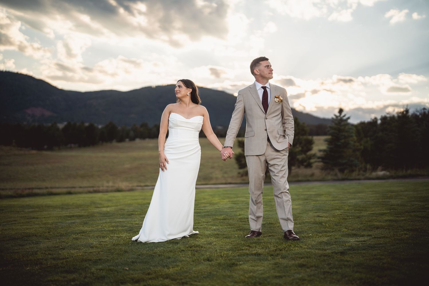 A wedding couple strolling hand in hand across an open field with mountains in the distance.