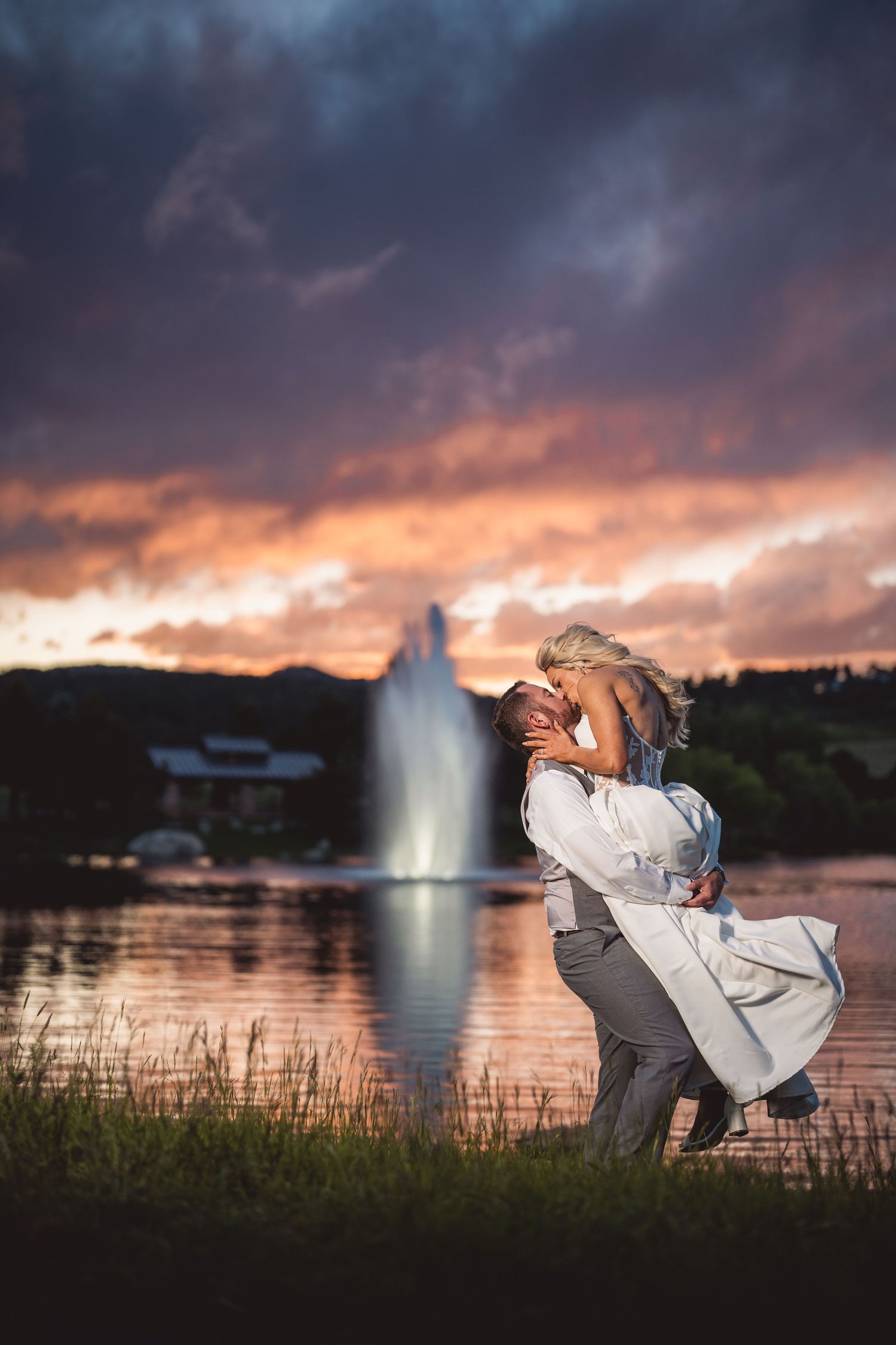 Couple sharing a romantic kiss at sunset by a fountain, bride in white gown lifted by groom under dramatic purple sky.