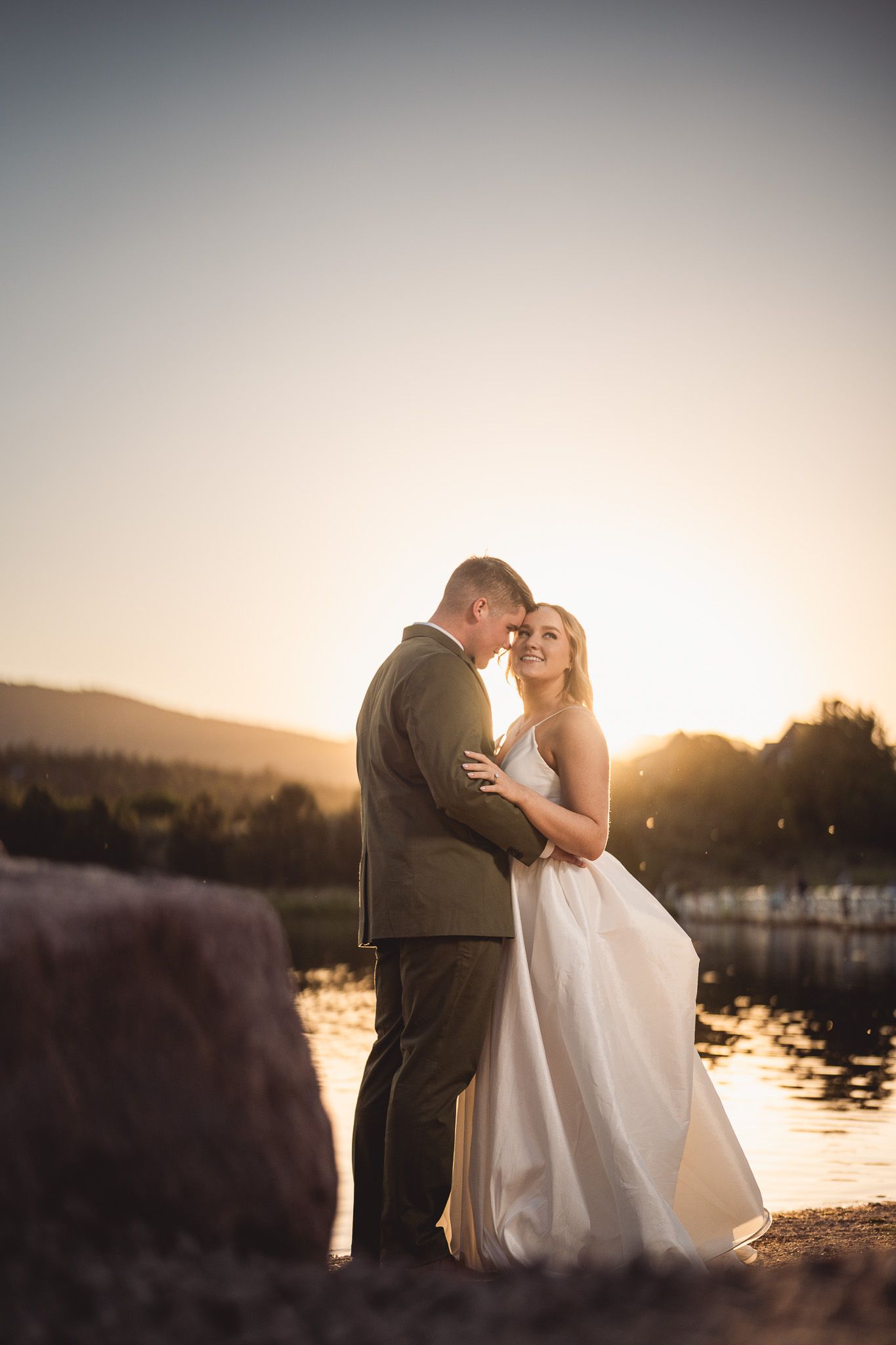Bride and groom embracing at golden sunset by a lake, bride in white gown, groom in olive suit.