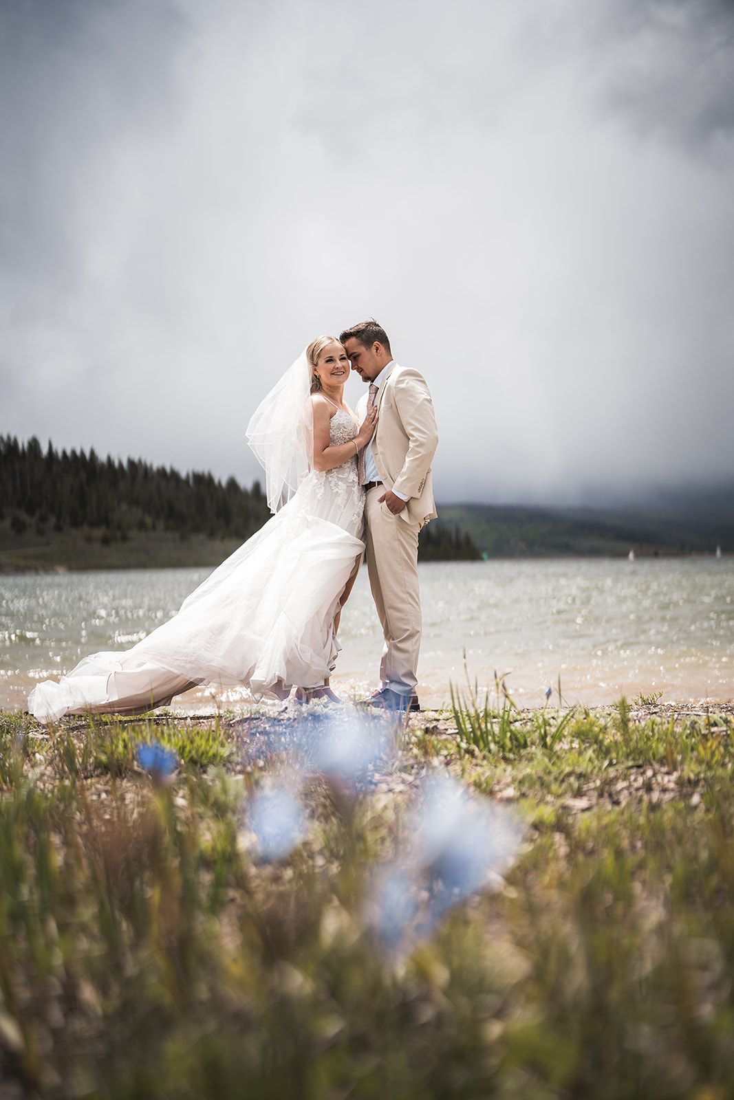 Bride and groom embrace by a mountain lake, she in a white gown with veil, he in a beige suit, under cloudy skies.