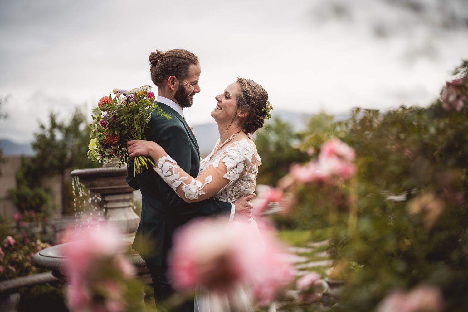 Bride and groom embrace in a rose garden, she holds a bouquet, wearing lace gown, he in dark suit.