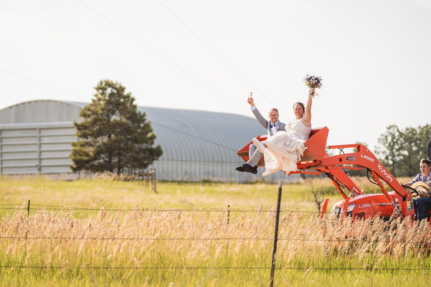 Bride and groom celebrate on a cherry picker lift at a farm wedding venue with a metal barn in the background.