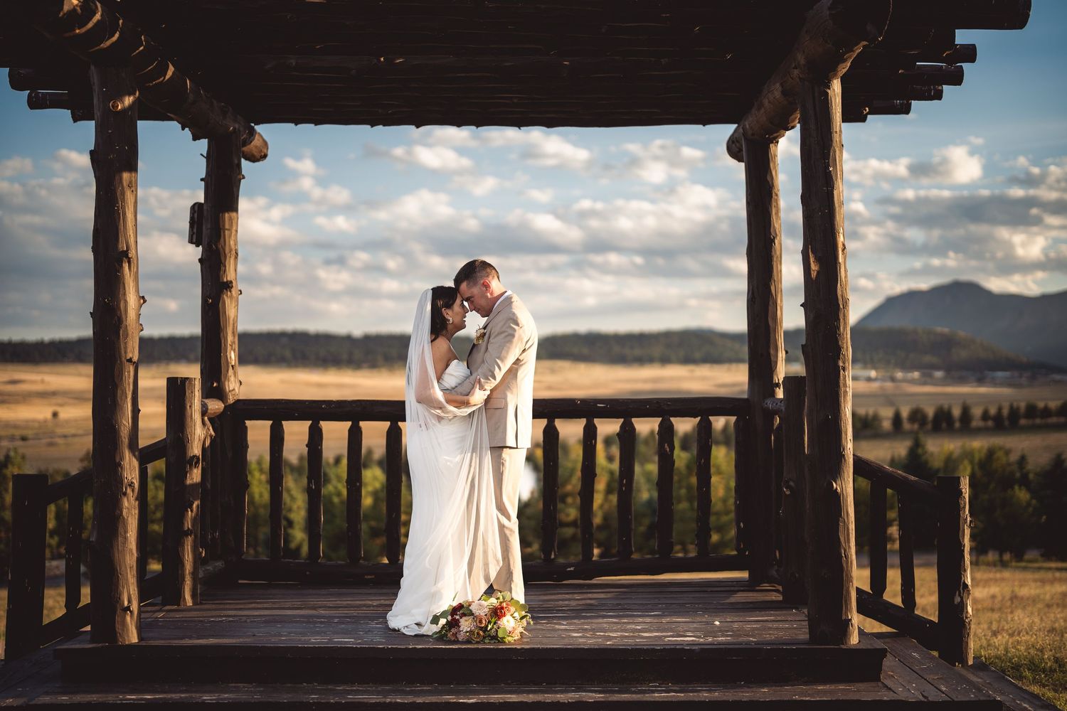 Bride and groom share a romantic kiss under a rustic wooden gazebo overlooking a scenic mountain valley.