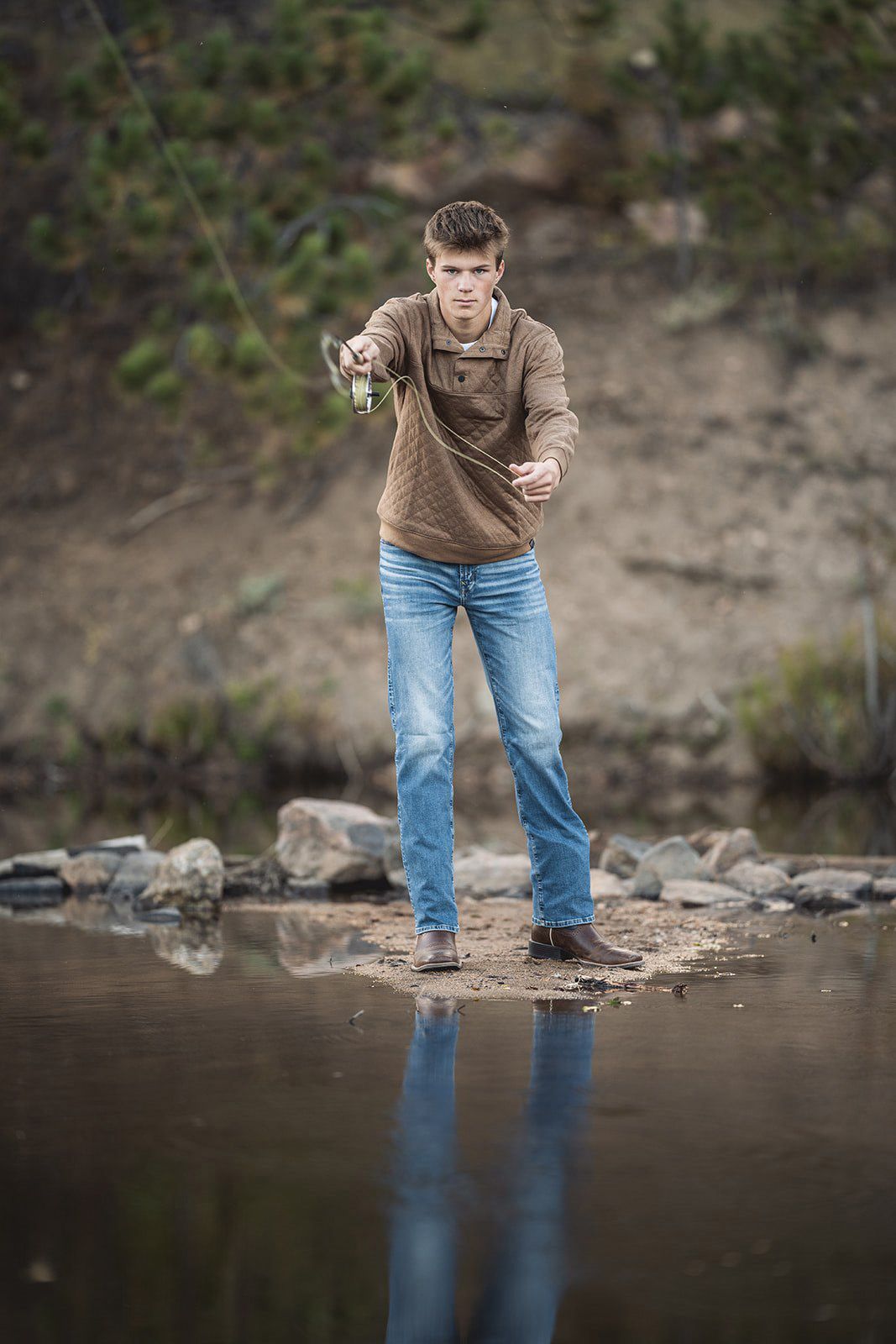 Young man fishing at a rocky riverbank, holding a fishing rod and lure while standing near calm water.