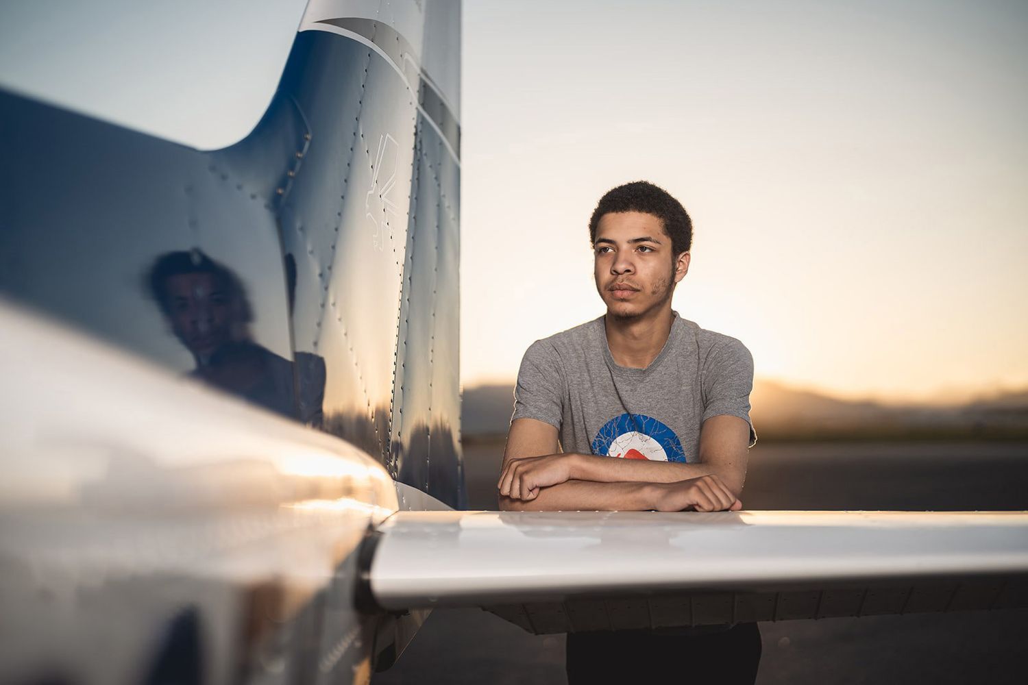 Young man resting arms on airplane wing at dusk, with his reflection visible on the aircraft surface.