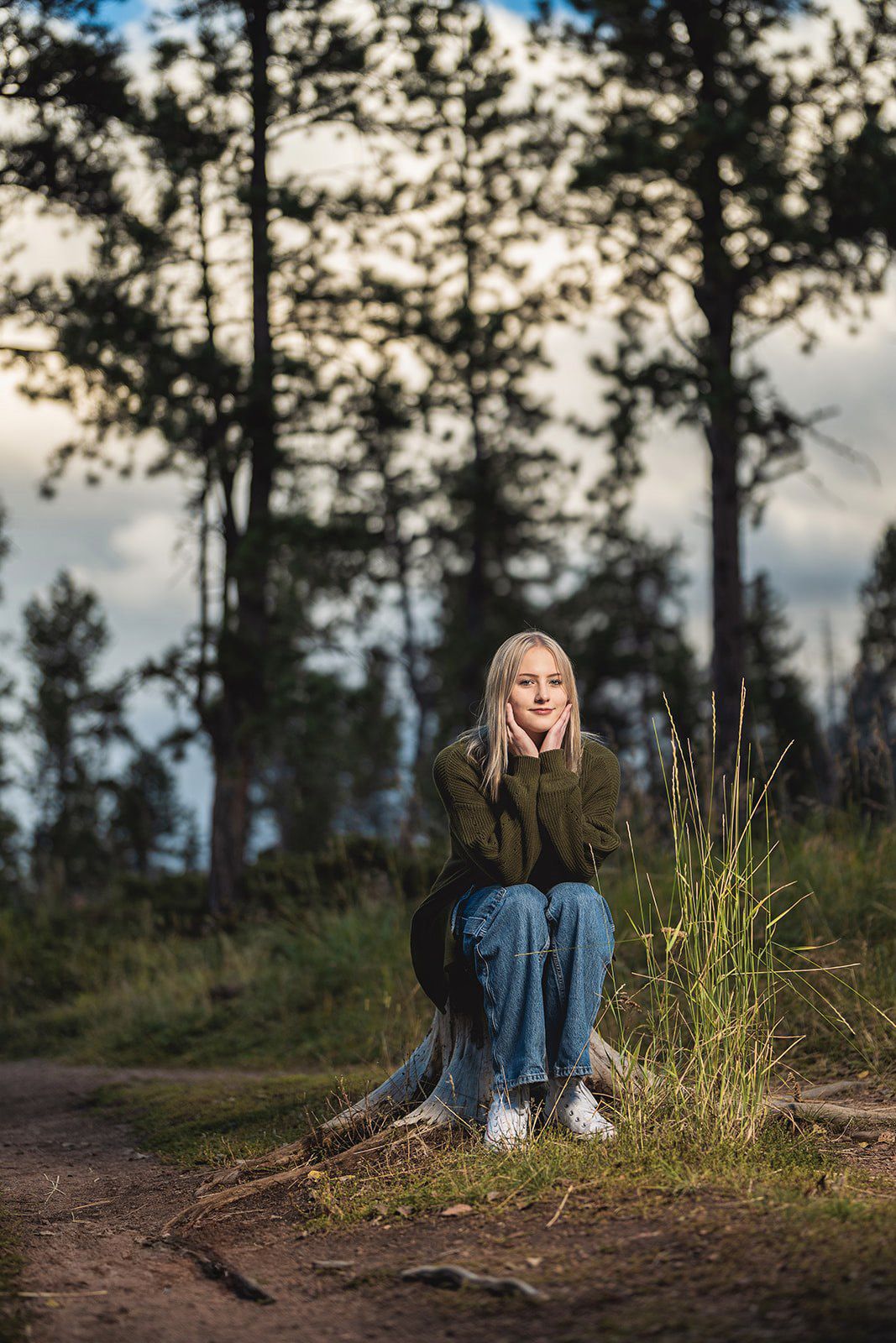 Blonde teen girl sitting on a tree stump in a forest clearing at dusk, hands on cheeks.