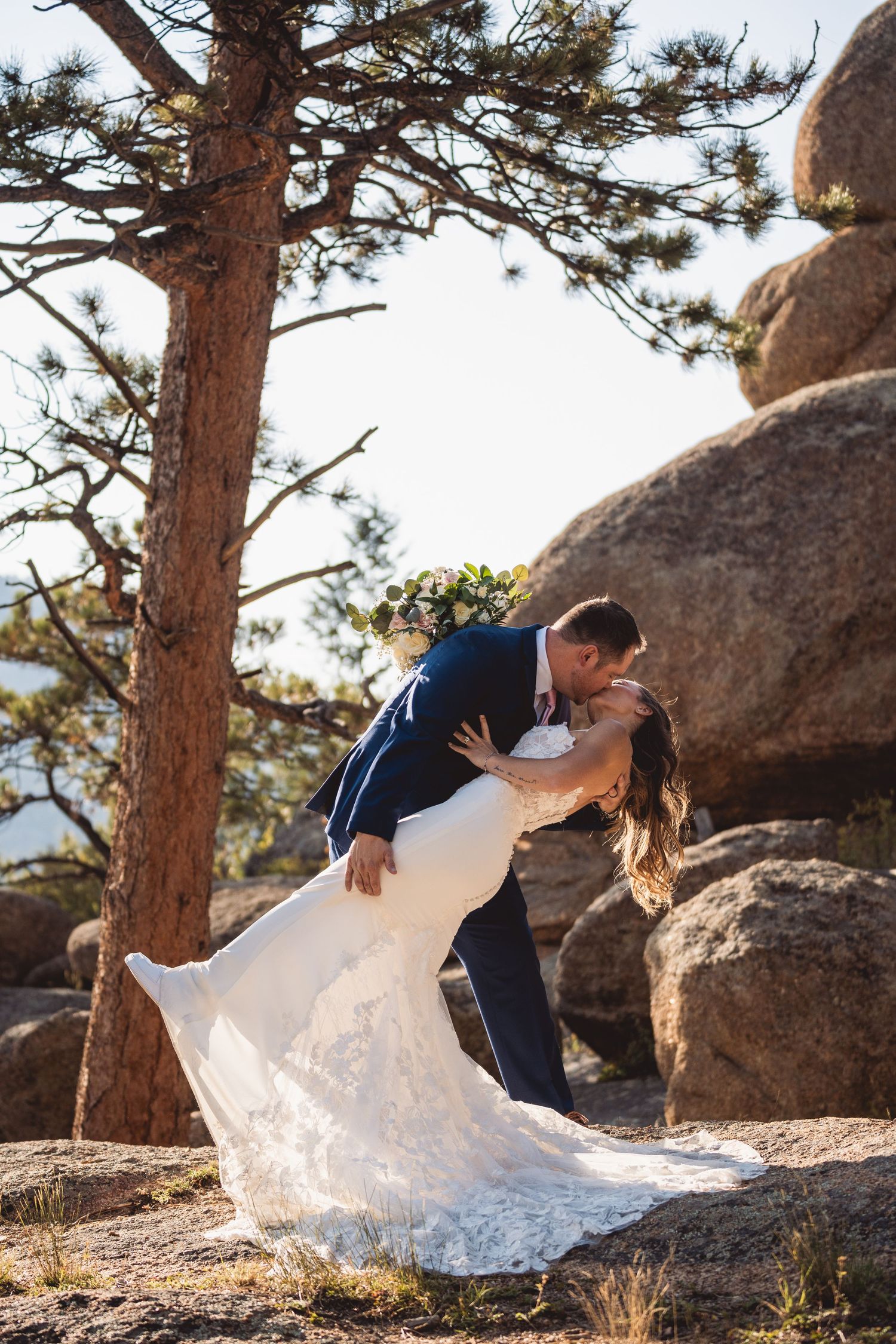 Bride and groom share a romantic dip kiss among rocky boulders and pine trees in an outdoor wedding photo.