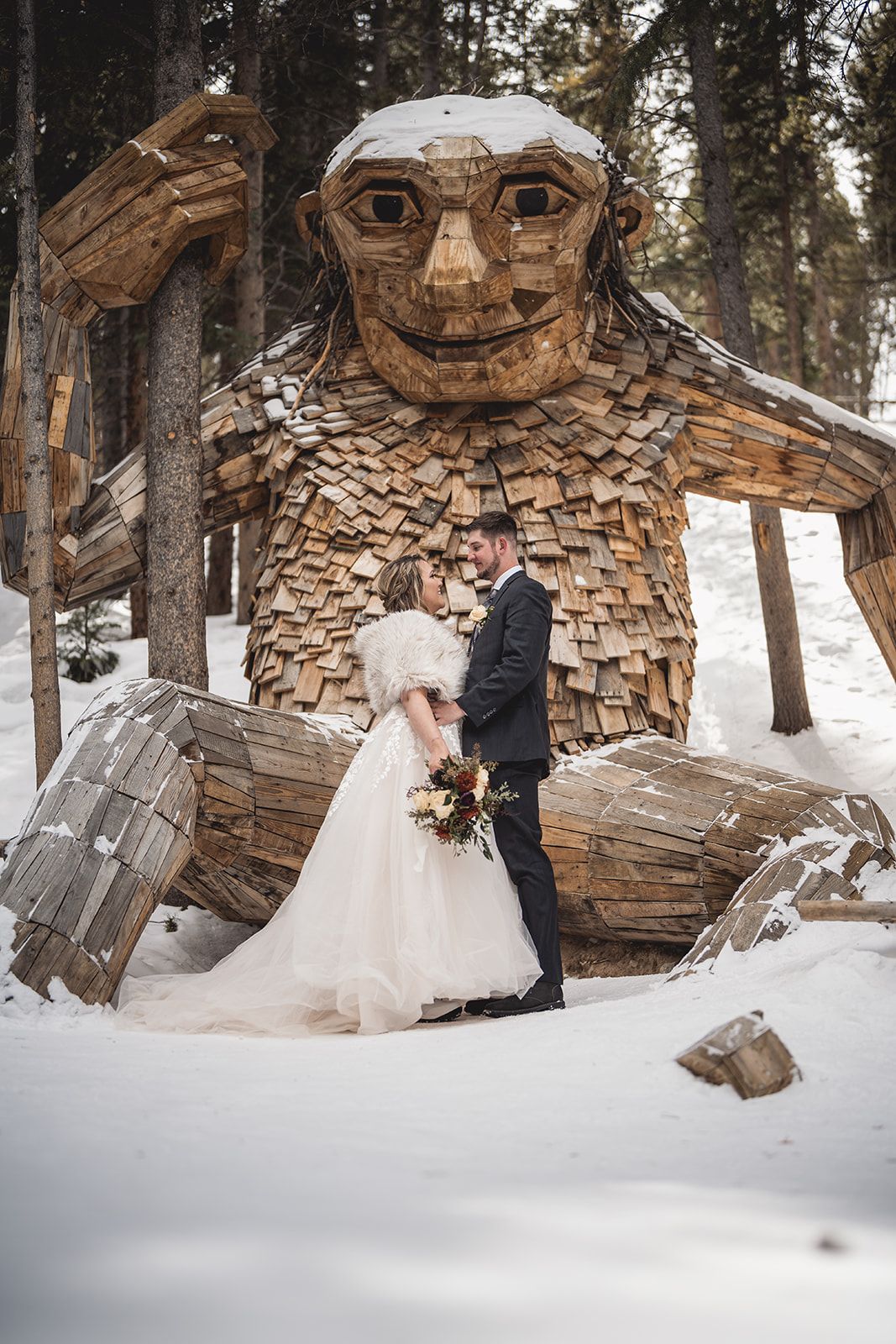 Bride and groom embracing in snow before a large wooden troll sculpture in a winter forest setting.