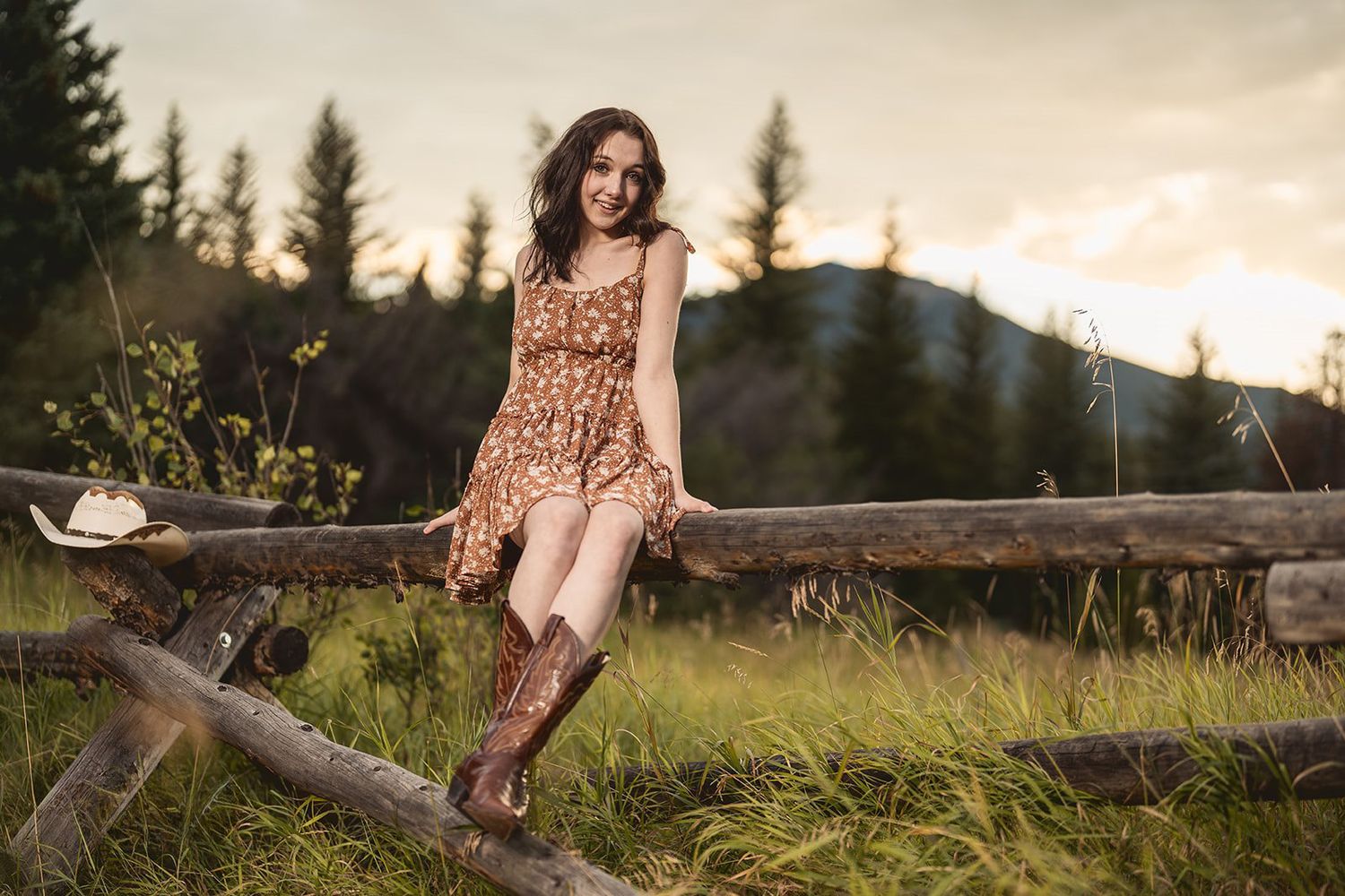 Smiling brunette in floral dress and cowboy boots sits on wooden fence at dusk.
