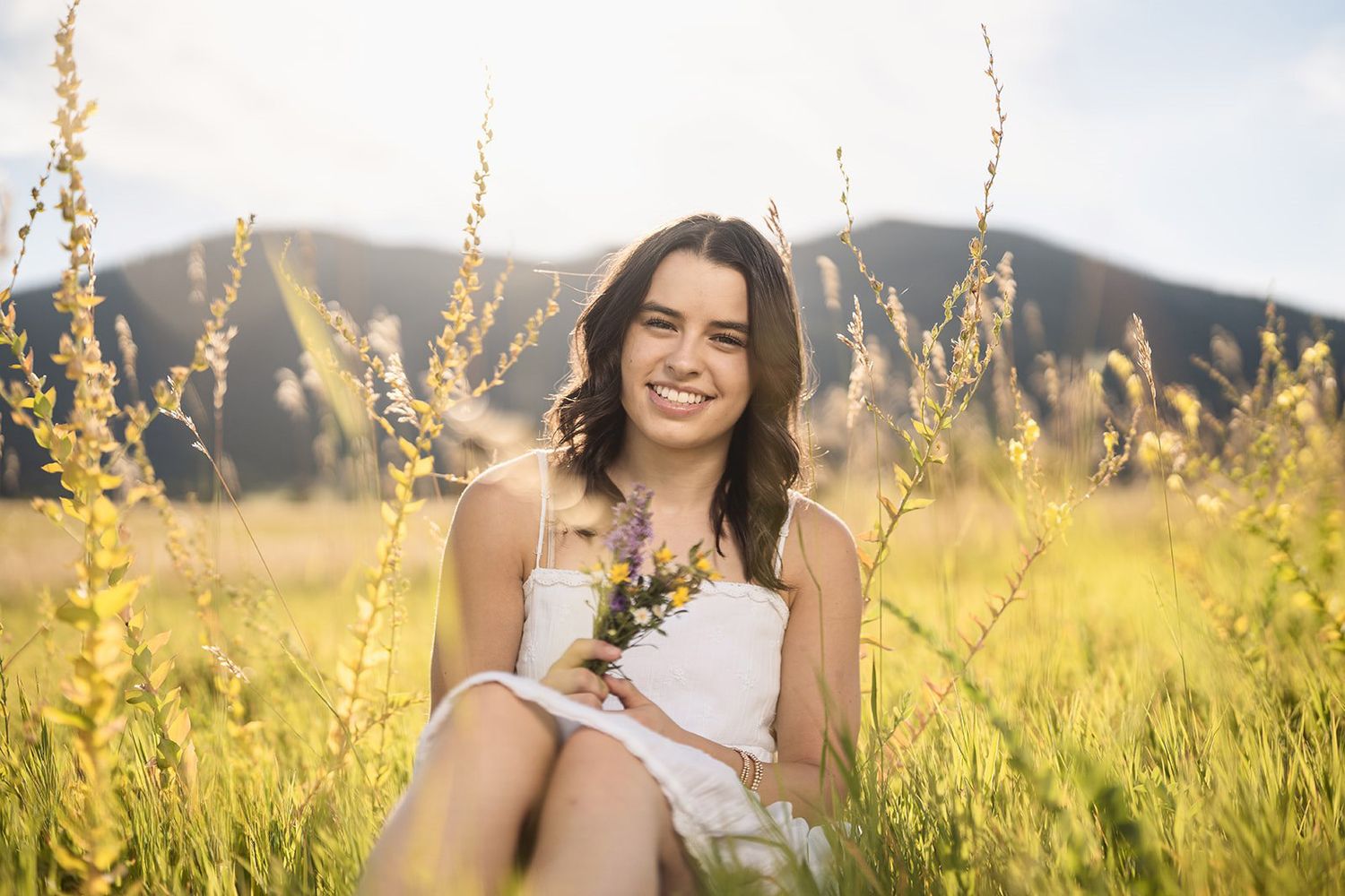Young woman in white dress sitting in sunny meadow with wildflowers and mountains, smiling and holding a bouquet.