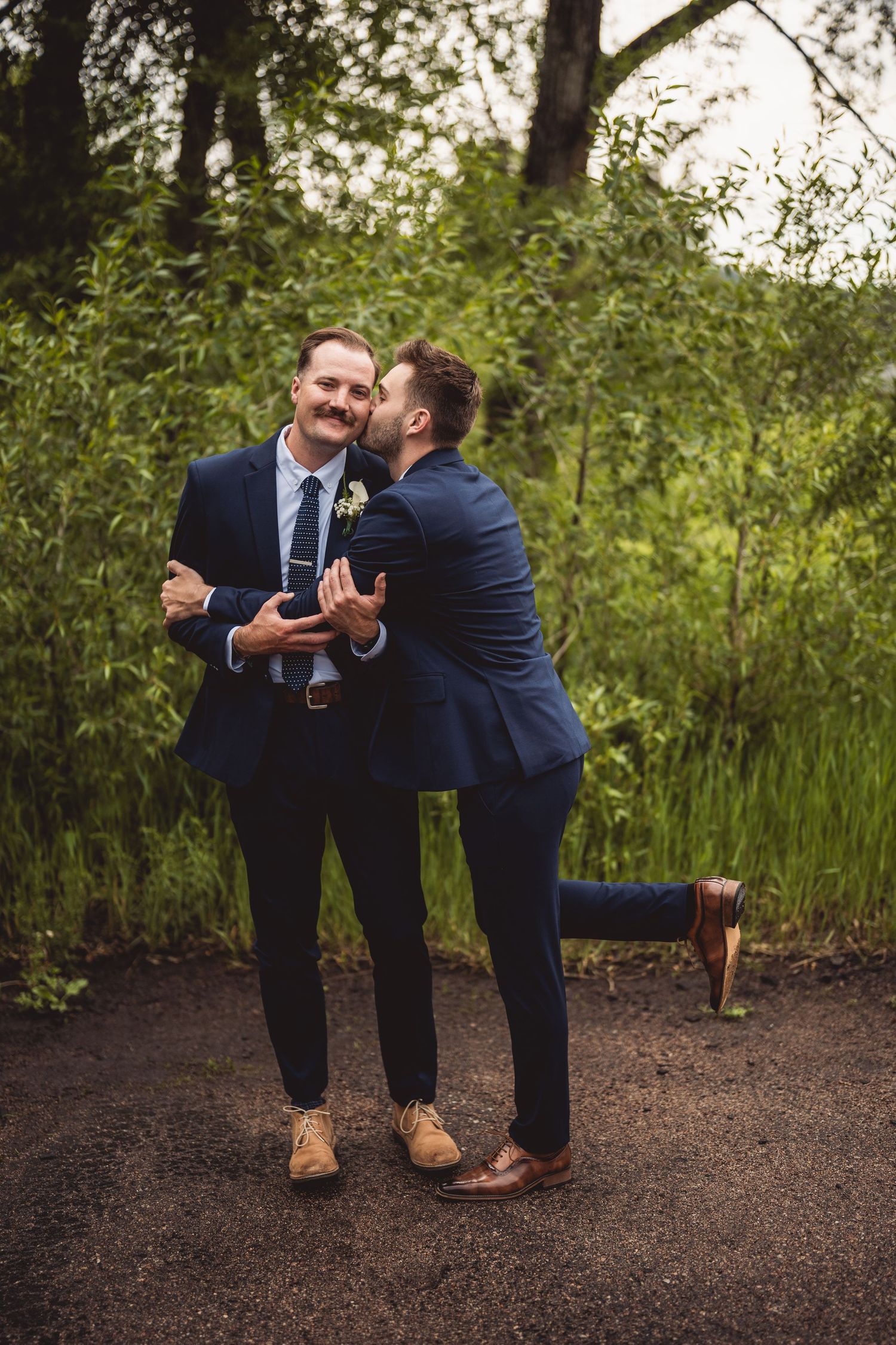 Two people in navy suits playfully interact on a dirt path surrounded by green foliage.