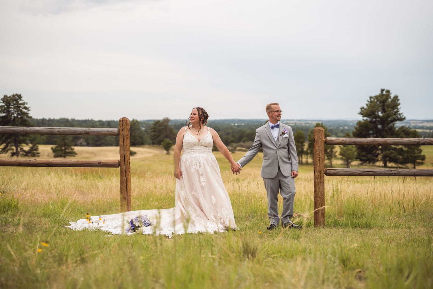 Wedding couple and attendants pose along rustic wooden fence in scenic meadow.