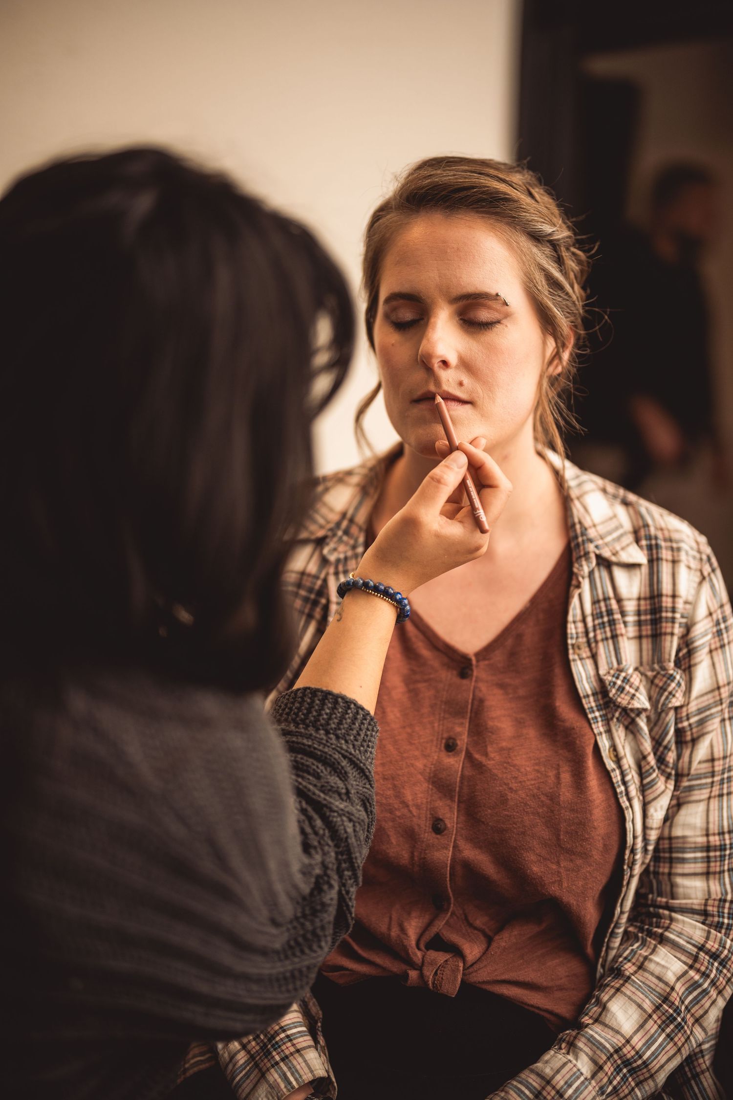 Behind the scenes makeup application in a dimly lit room with plaid shirt.