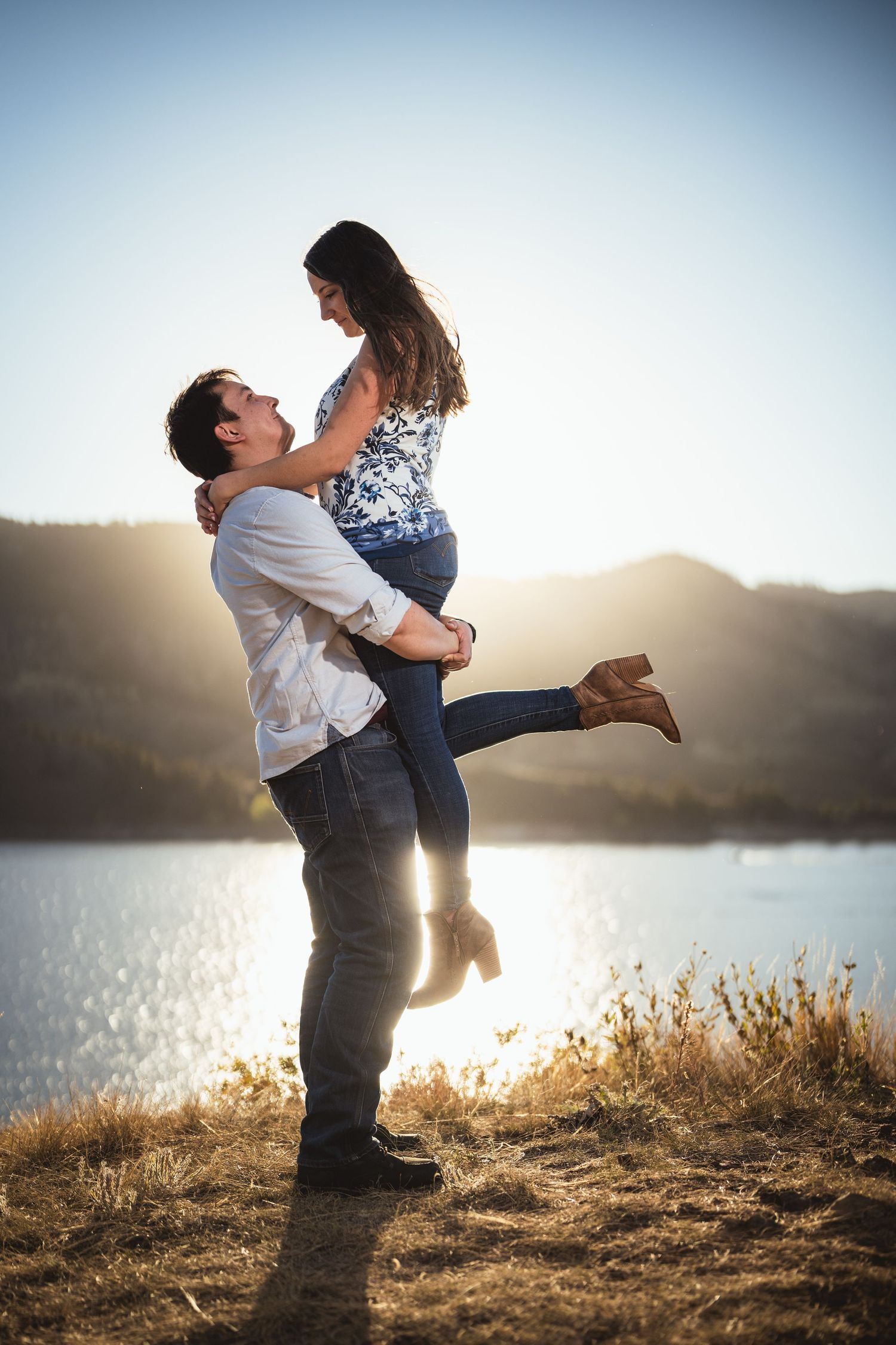 Couple shares a playful lifting embrace against a sunset lake backdrop.