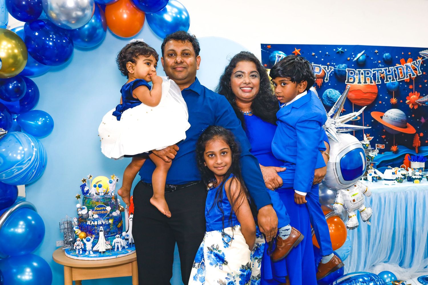 A family of five posing for a photo wearing blue themed outfits at a kid's birthday party. A cake is in the background and decorations.