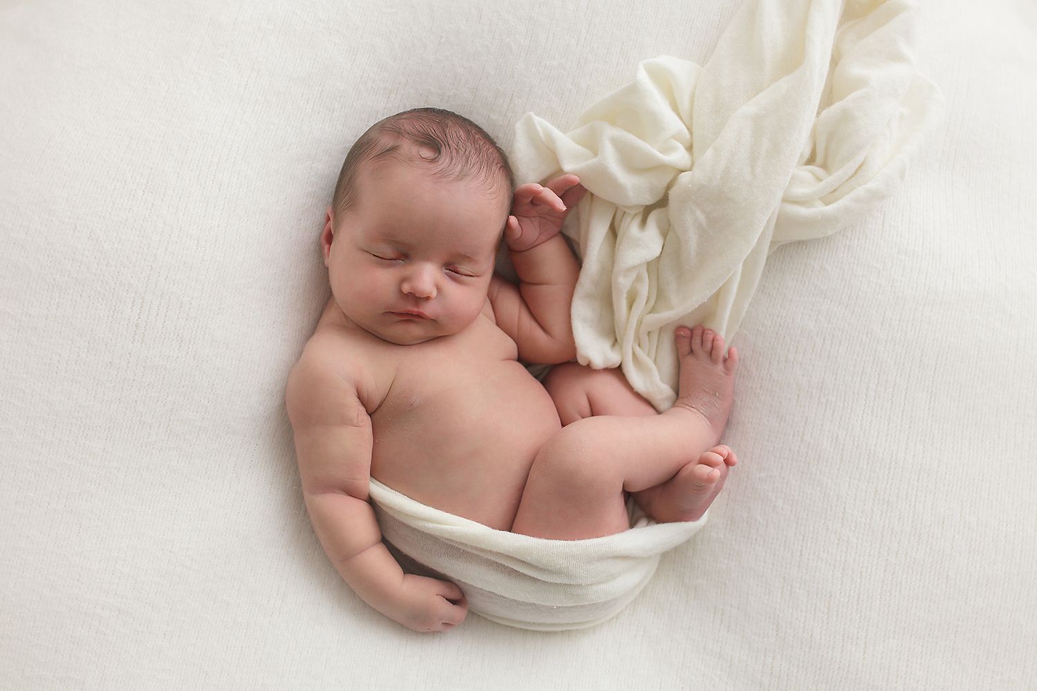 Sleeping newborn baby wrapped in soft white fabric against light background.