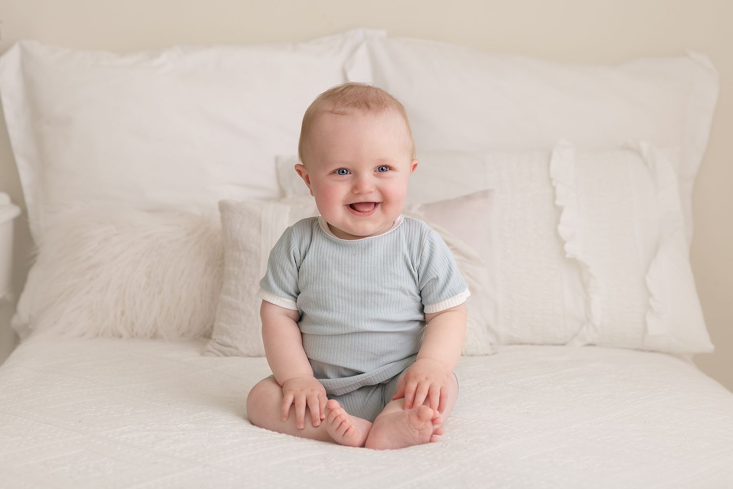 Baby wearing light blue shirt sits on white bedding during portrait session.