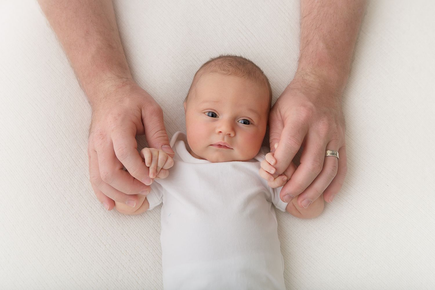 Baby in white onesie lying on white background with hands framing their face.