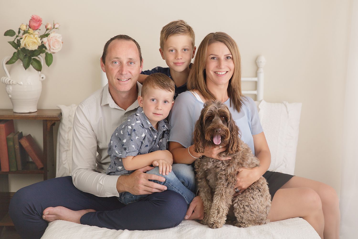 Family portrait on white couch with chocolate labradoodle dog and pink roses in background.