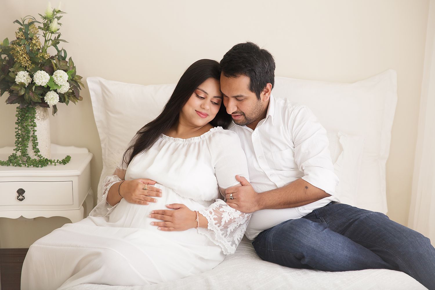 Expectant couple in white sharing intimate moment on bed with floral decor.
