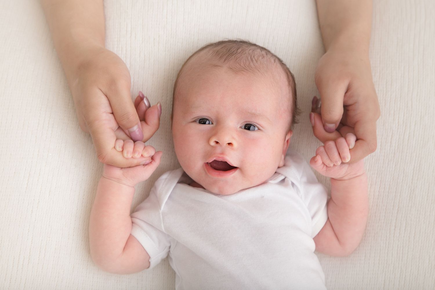 Smiling baby in white onesie lying on white background with hands being held.