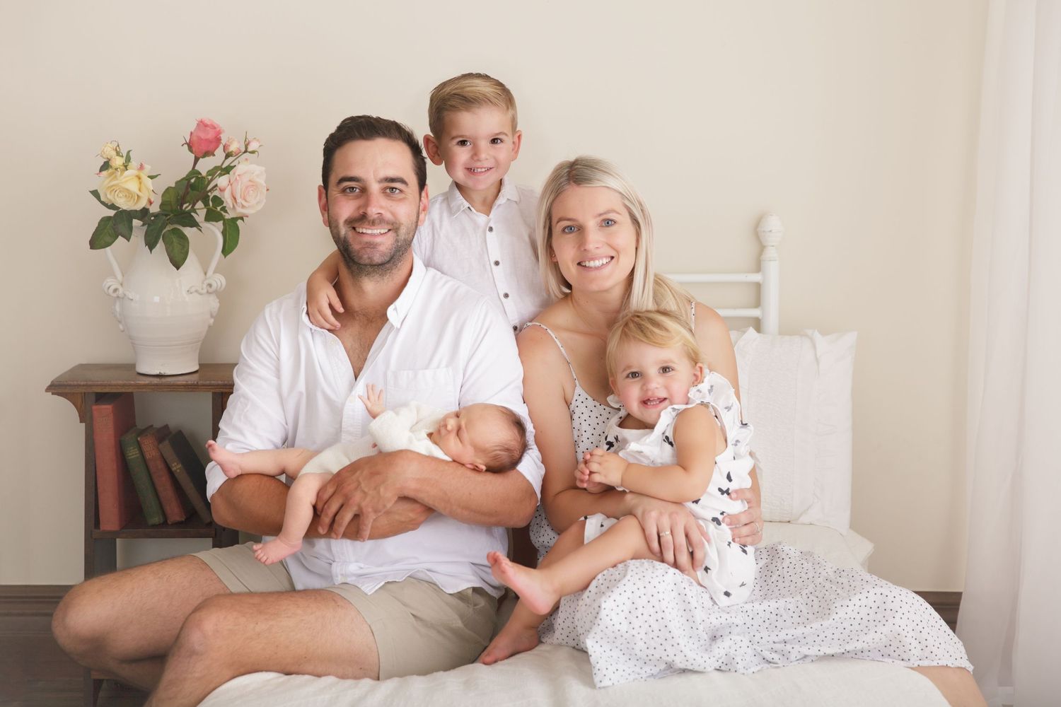 Family portrait with newborn baby and toddler on white bed with pink roses.