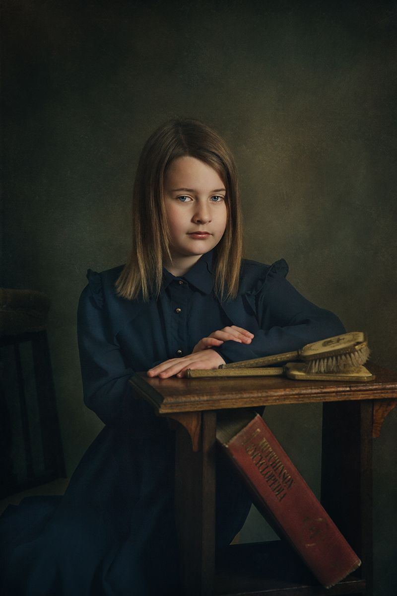 Artistic portrait in dark tones showing someone at an antique wooden desk in navy blue attire.