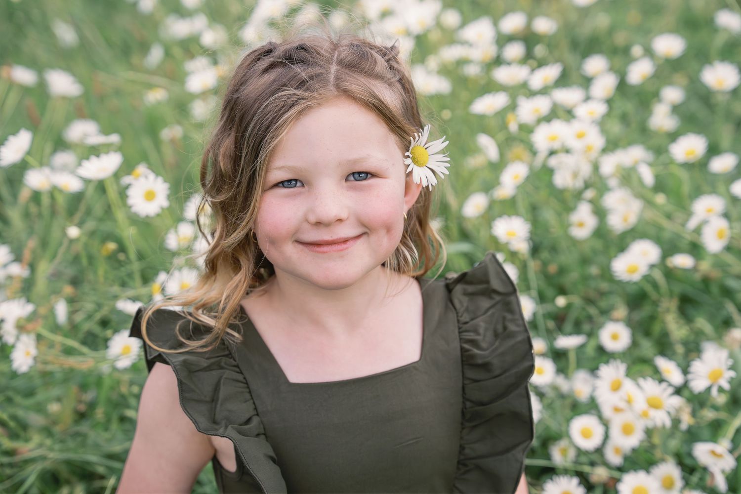 A child wearing a green dress poses in a field of blooming white daisies on a sunny summer day.