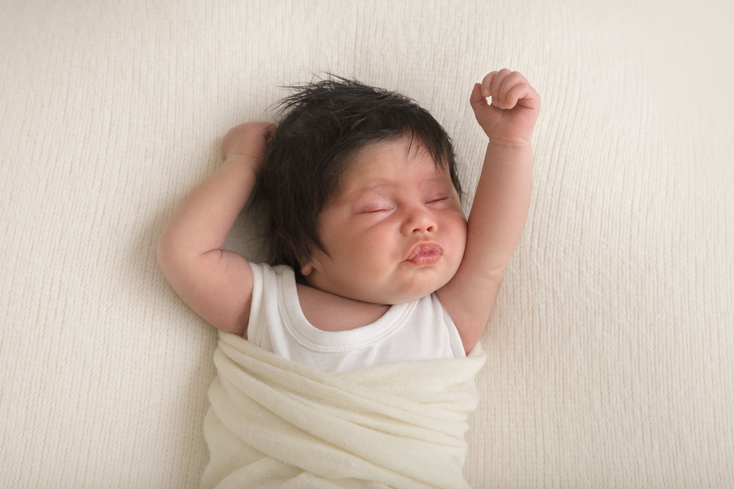 Sleeping baby with dark hair wrapped in cream colored fabric on light background.