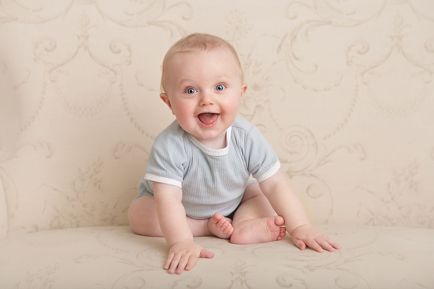 Adorable baby in grey onesie sitting on white backdrop with joyful expression.