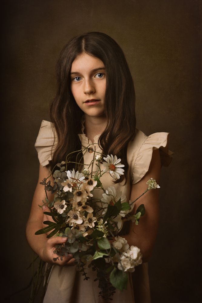 Young girl in vintage dress holding white and gold floral bouquet against dark backdrop.