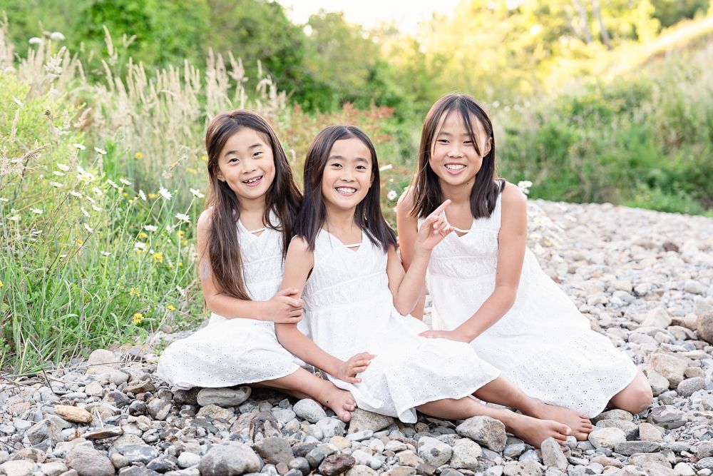 Three people in white dresses sit together on a rocky surface surrounded by green foliage.
